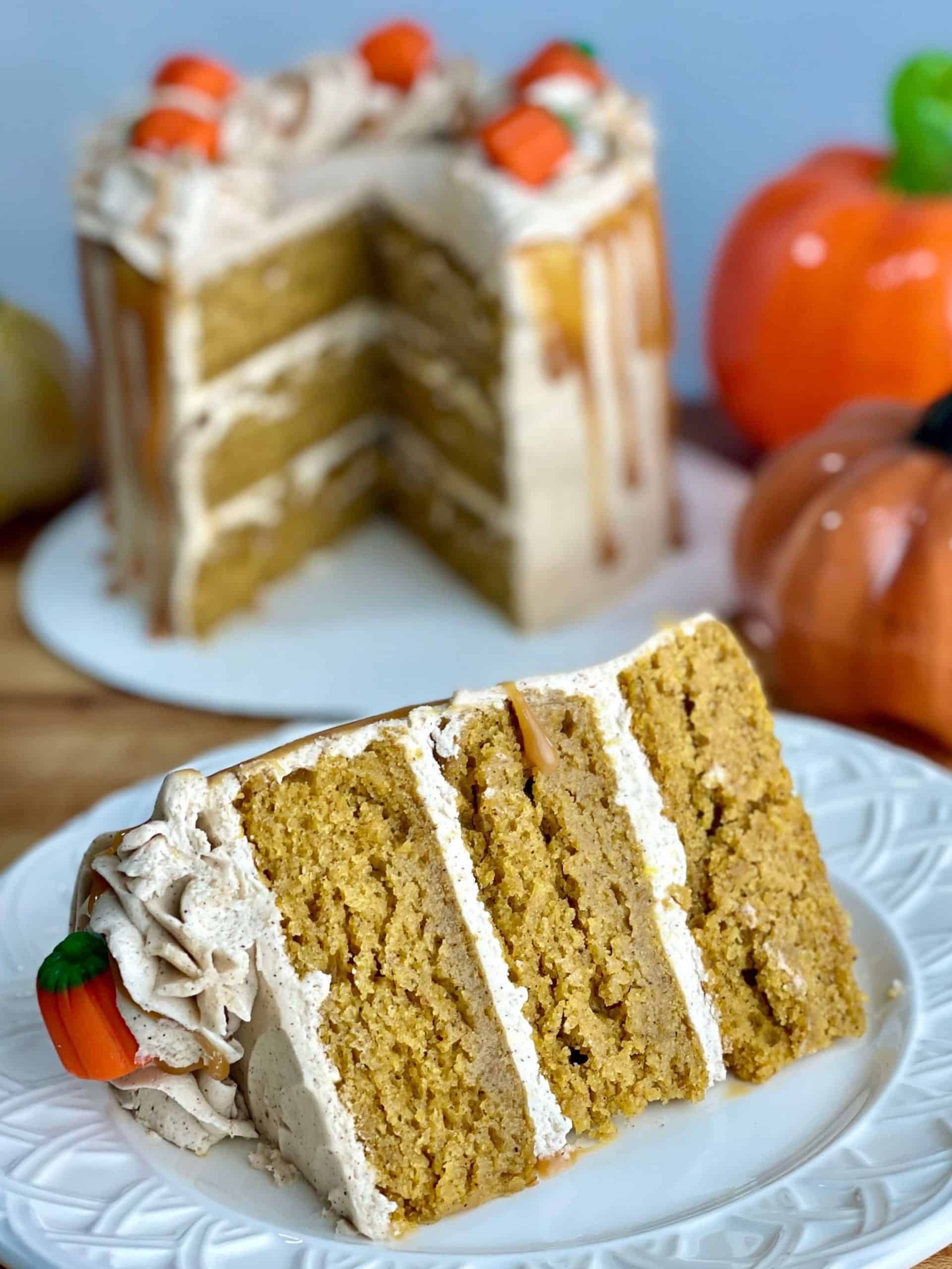 a closeup of a slice of three layer pumpkin spice cake that is visibly very moist with a cut layer cake in the background and some glass pumpkins
