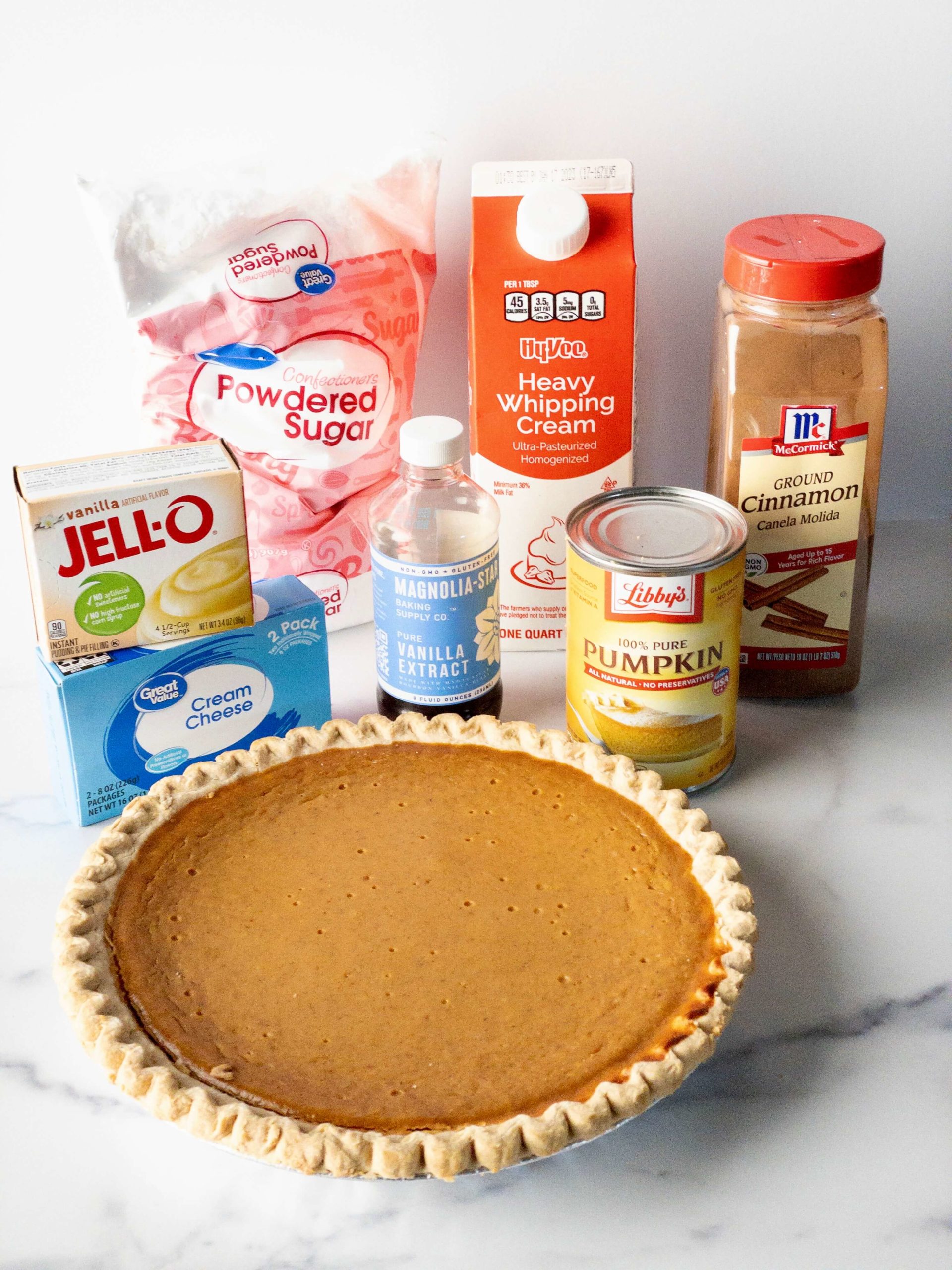 Pumpkin Cream pie ingredients on a table--a premade pumpkin pie, powdered sugar, cream cheese, heavy cream, cinnamon, canned pumpkin puree, pure vanilla extract, and vanilla instant pudding mix