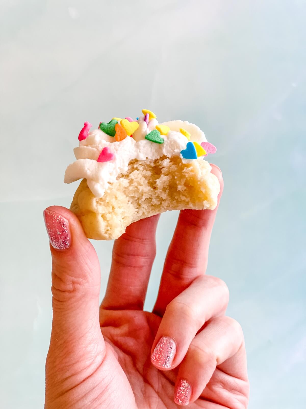 A hand holding a mini frosted sugar cookie with a bite taken out, showing the soft center