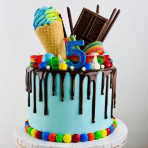 A blue cake with chocolate drip and colorful edible toppings
