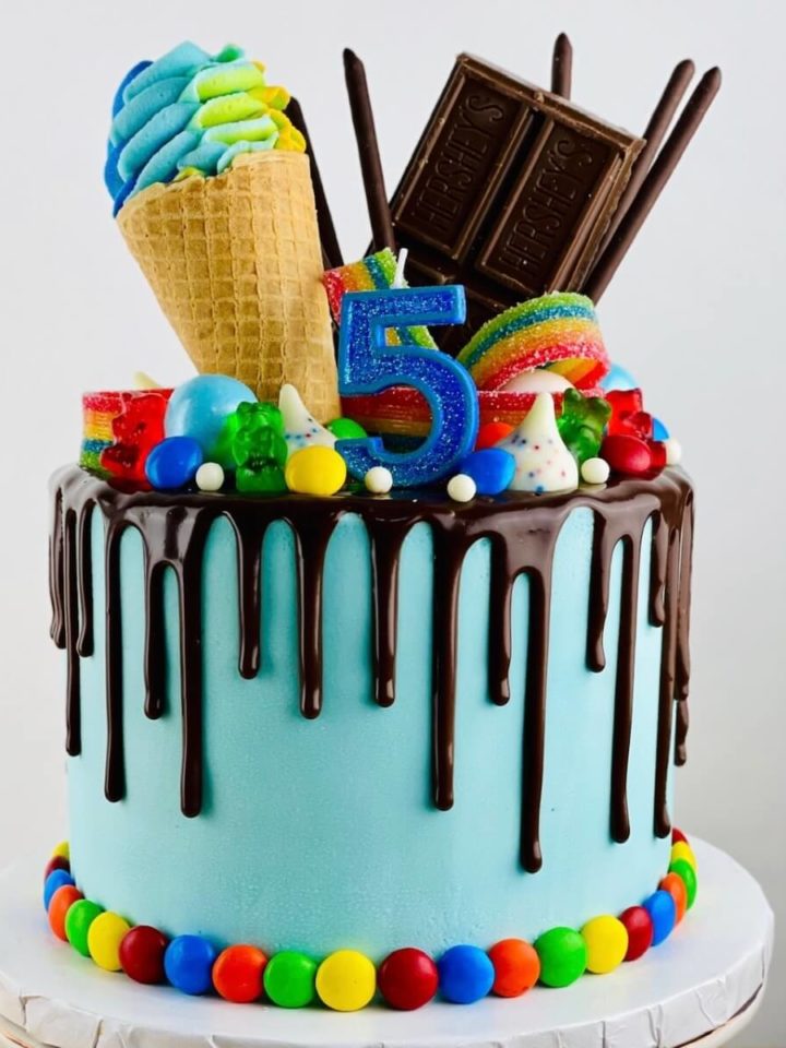 A blue cake with chocolate drip and colorful edible toppings