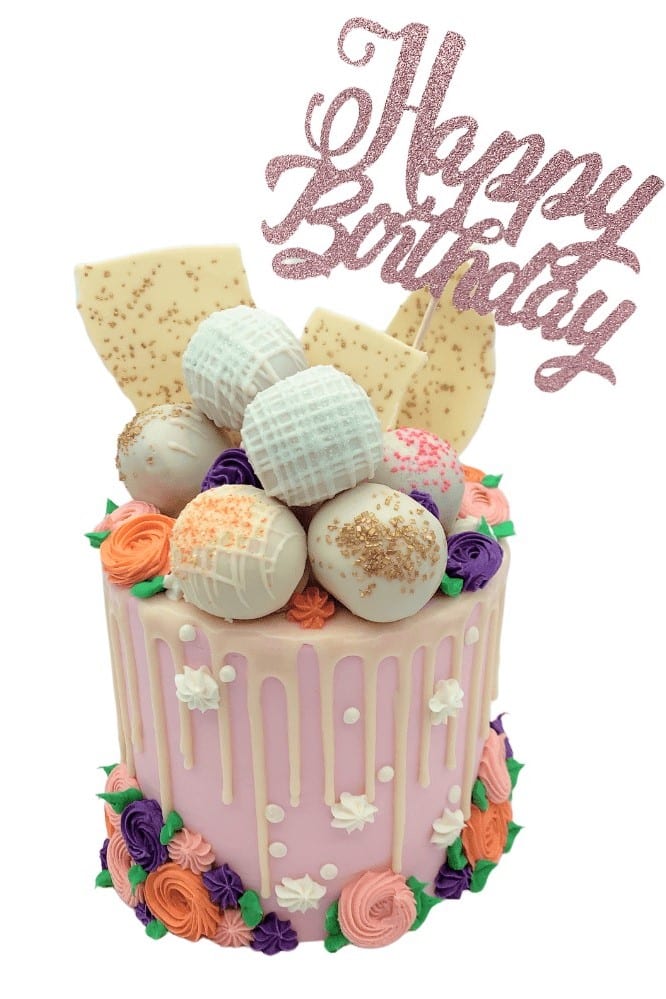 a pink drip cake with white ganache drizzle, cake truffles on top and a happy birthday banner