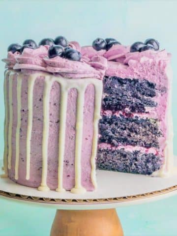 A blueberry cake on a pedestal with a slice taken out showing layers of moist blueberry cake, blueberry buttercream, and blueberry compote filling.