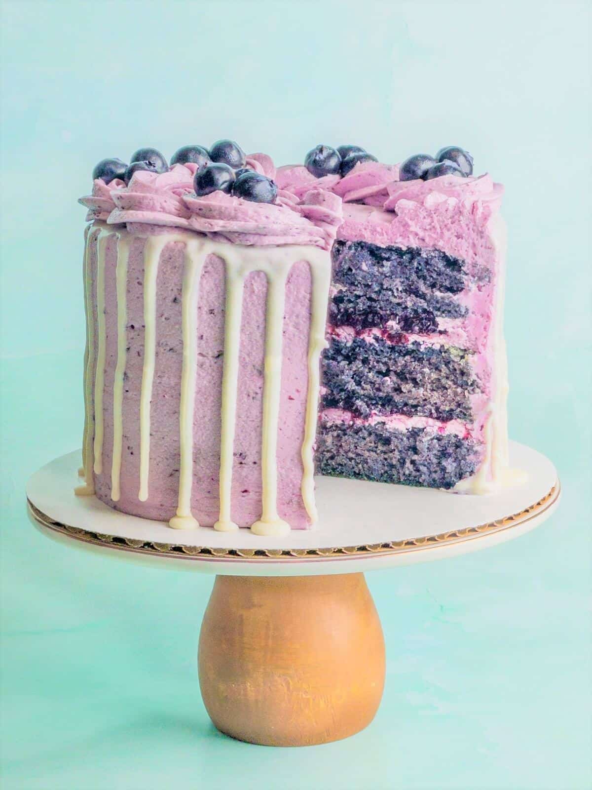 A beautiful blueberry cake on a cake pedestal cut open showing three layers of cake, blueberry filling and blueberry frosting.