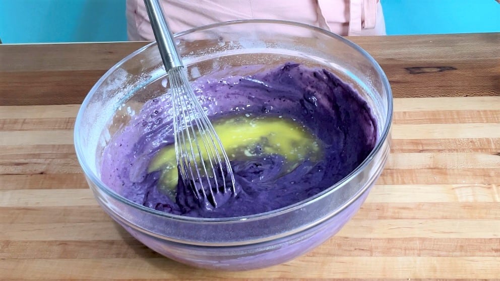 A bowl with blueberry cake batter being whisked together with melted butter.