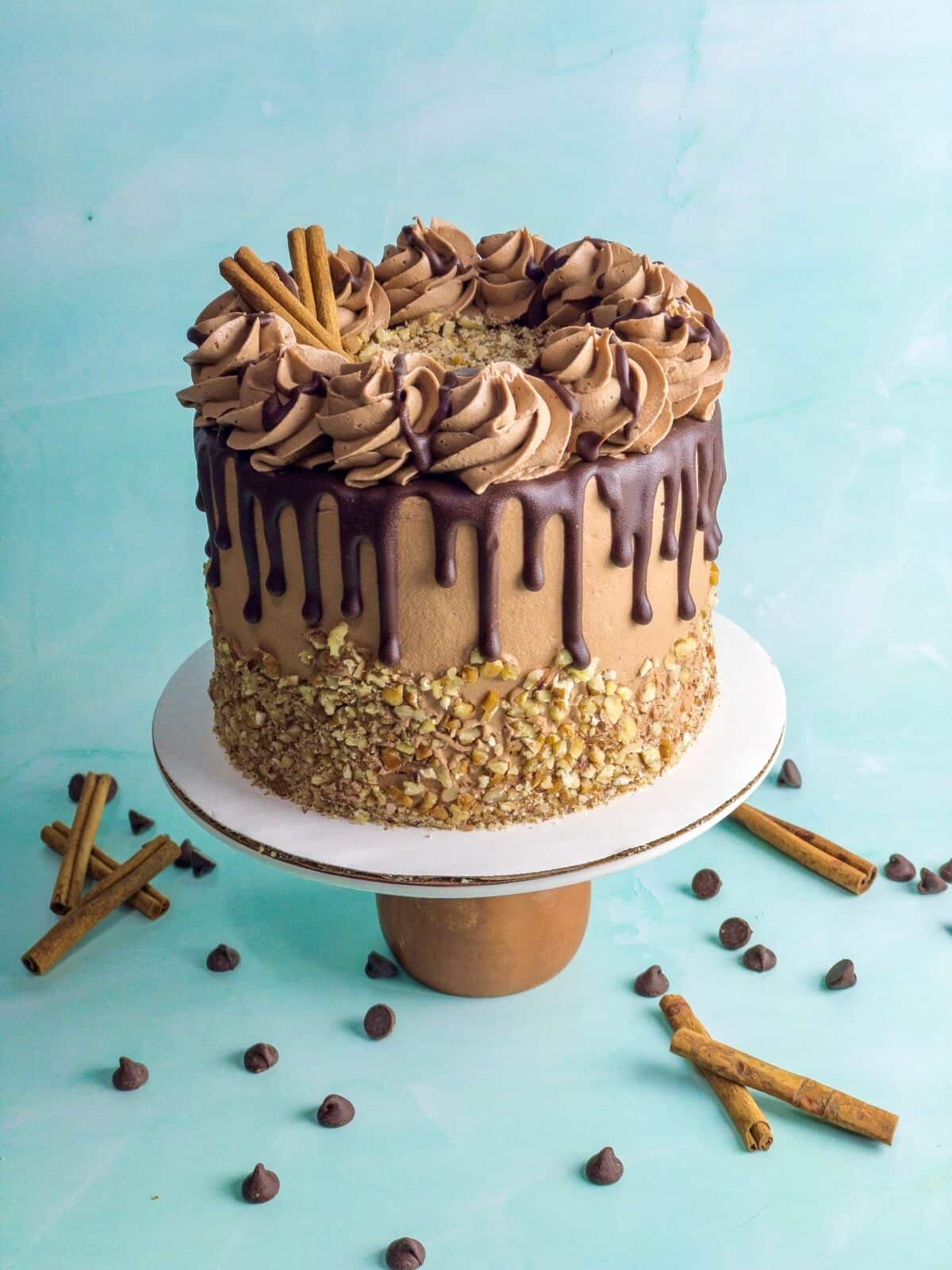 an image of an uncut chocolate cinnamon drip cake with pecans around the edges and chocolate ganache drizzle down the sides of the cake. On a cake pedistal with chocolate chips and cinnamon sticks on the table