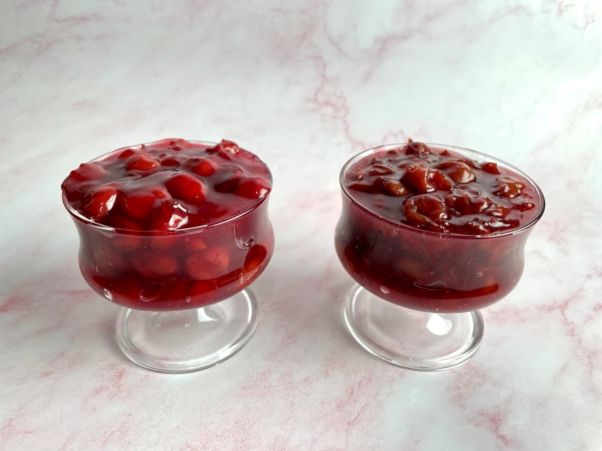 A dish of cherry filling made with frozen cherries that is bright red with round cherries. Next to it is a dish of cherry filling made with canned cherries that is darker in color and the cherries are less round