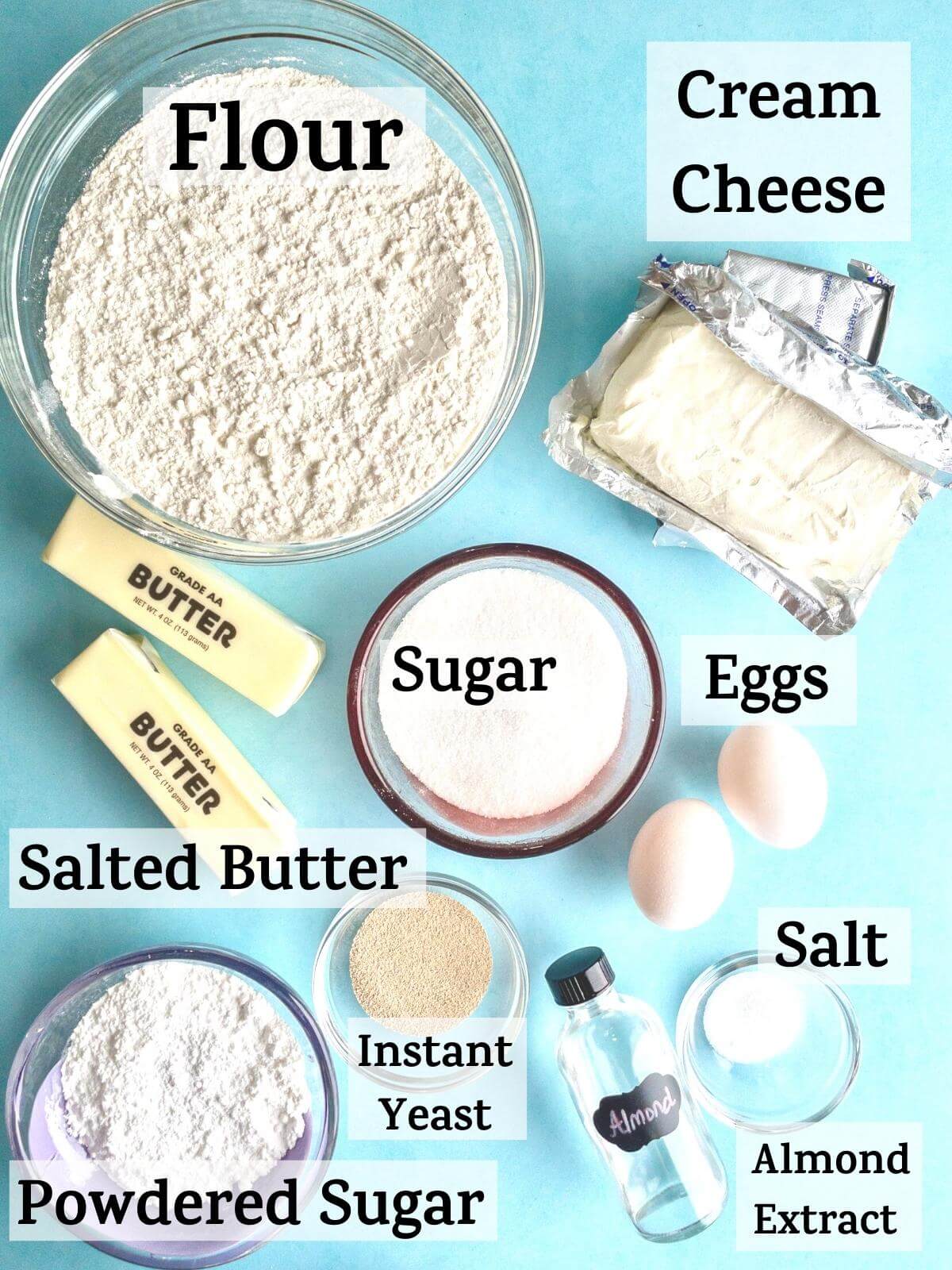 An overhead shot of cream cheese danish ingredients with the following text: flour, sugar, salted butter, eggs, salt, instant yeast, cream cheese, powdered sugar, almond extract.