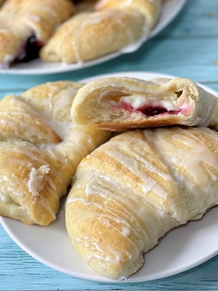 A closeup of soft cream cheese danish pastries, one open with the cream cheese and cherry filling showing on the inside.