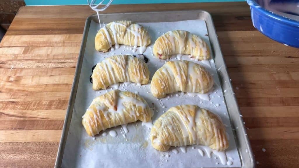 A sheet pan of baked danishes, half glazed and one being drizzled with glaze.