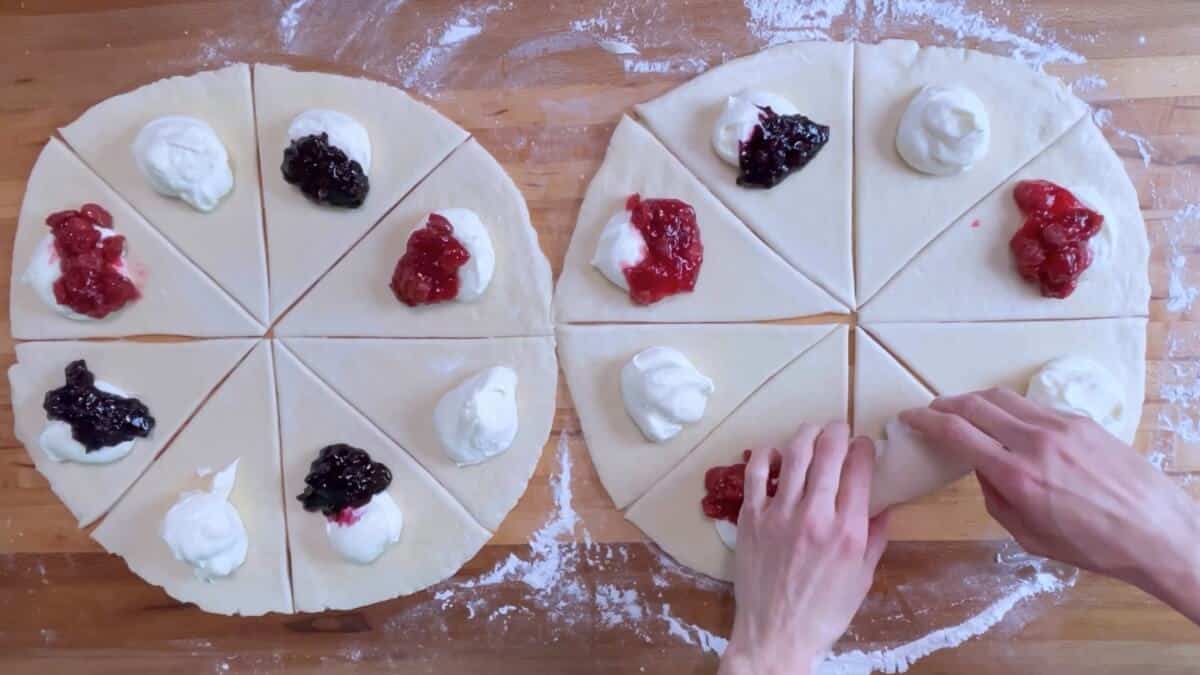 Two circles of rolled out danish dough cut into 8 triangles each, topped with cream cheese filling, cherry filling, and blueberry filling.