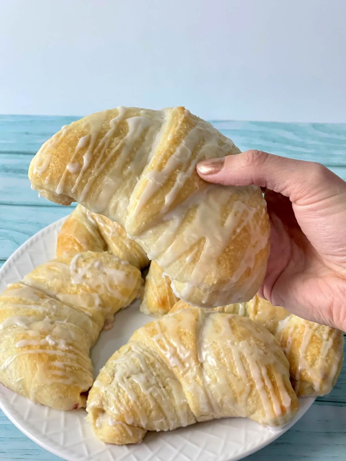 A picture with a hand holding a large danish with cream cheese filling, with a plate of danish drizzled with a powdered sugar glaze in the background.