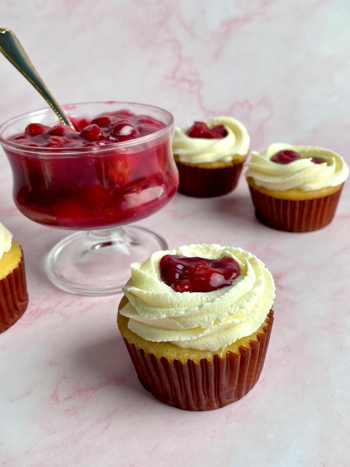 several vanilla cupakes with a dollop of homemade cherry cake filling on top of each cupcake, and a bowl of cherry filling in the background