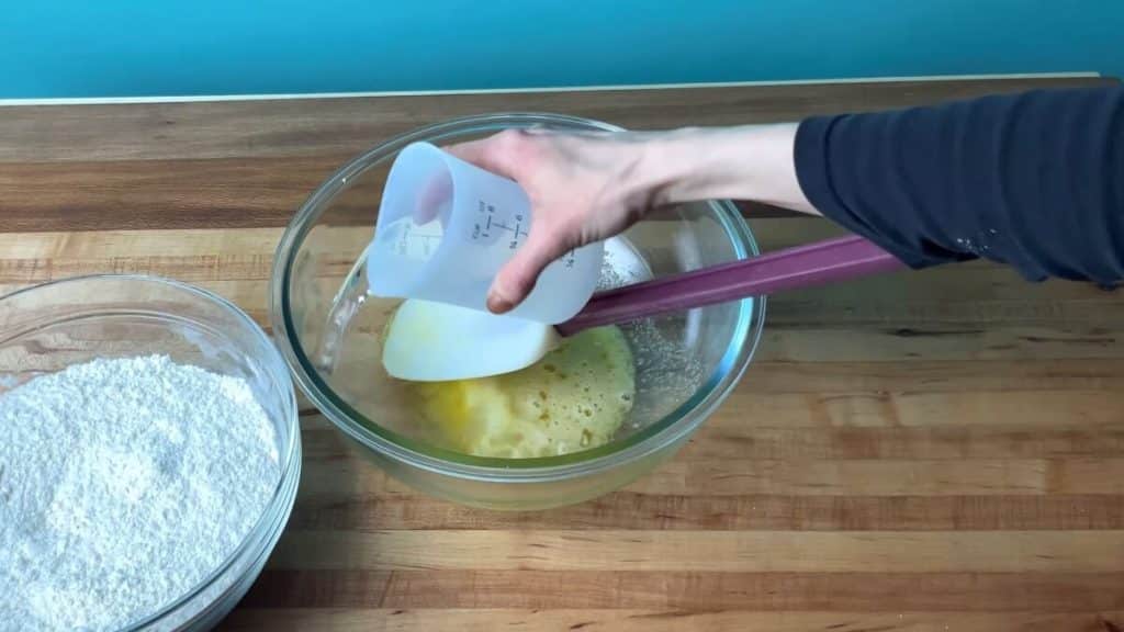 A measuring cup of water being poured over a bowl containing wet ingredients, and a second bowl of dry ingredients.