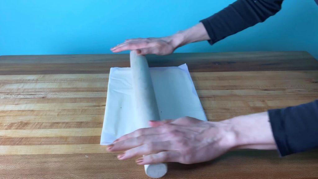 A folded piece of parchment paper containing butter being rolled smooth with a rolling pin.