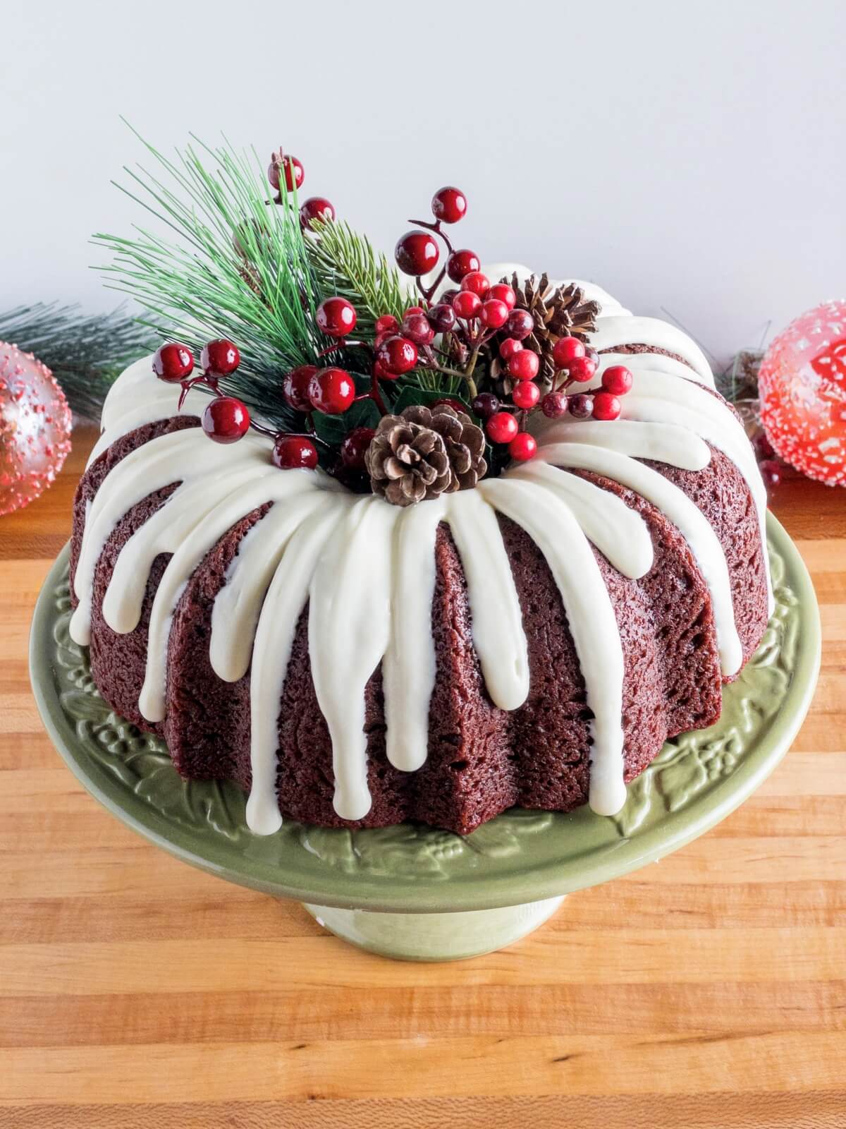 A Christmas Bundt Cake with red velvet cake, cream cheese glaze drizzle, and holly, berries and pine cones decorating the cake