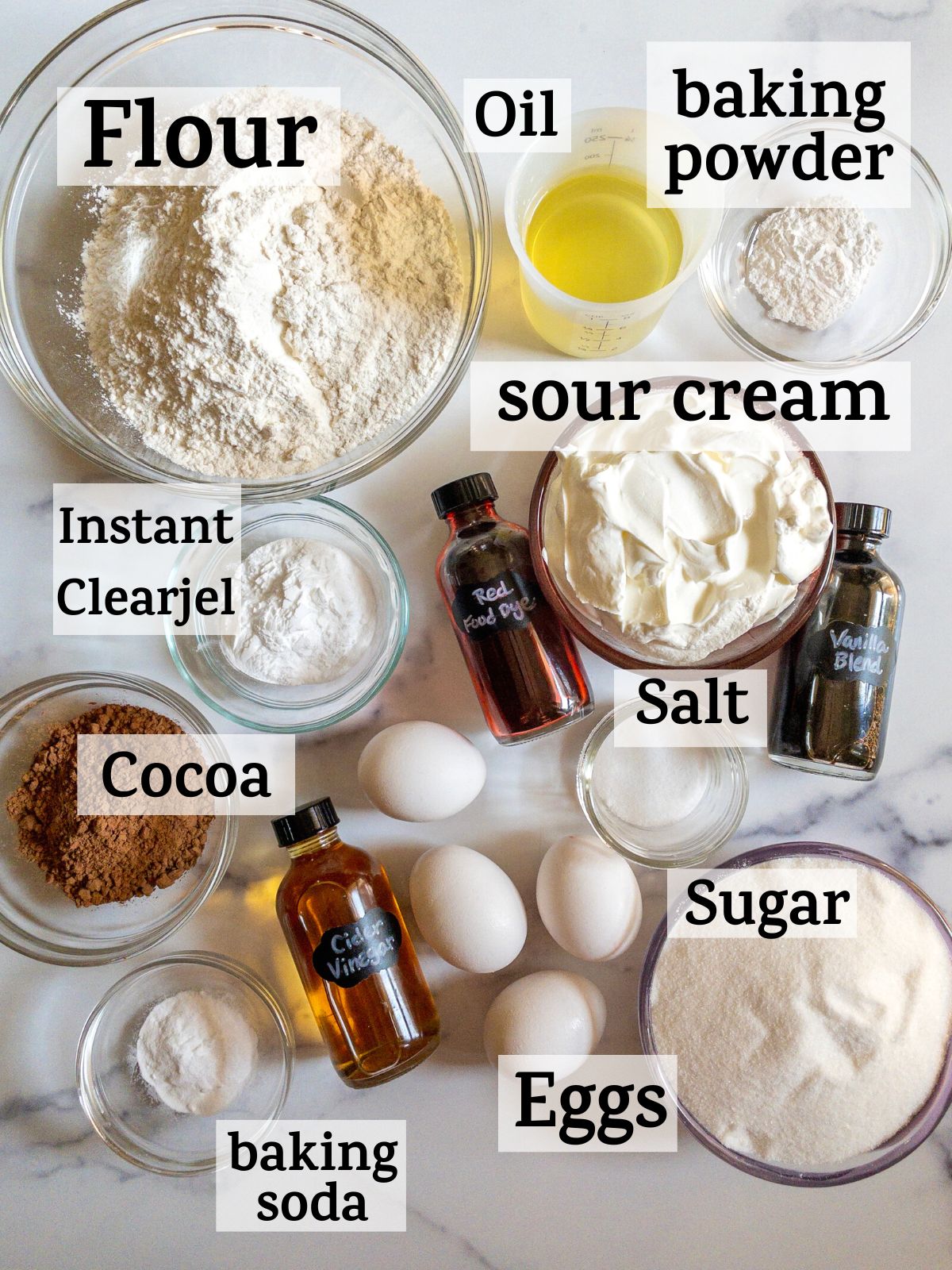 An overhead shot of red velvet cake ingredients, with the text "flour, sugar, instant clearjel, cocoa, vanilla blend, red food dye, cider vinegar, eggs, salt, baking powder, baking soda, sour cream, oil.