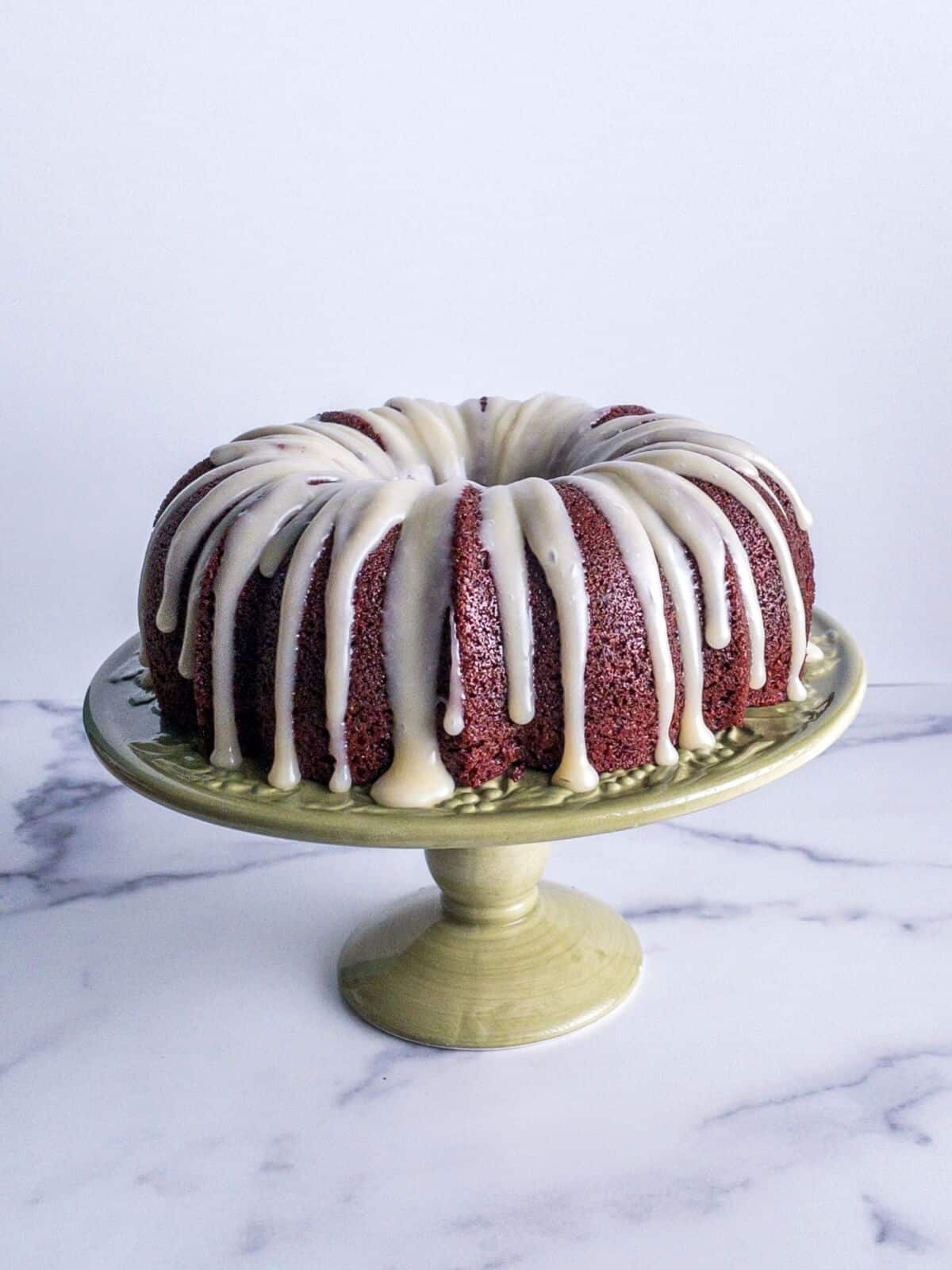 A red velvet bundt cake on a green cake pedestal with cream cheese glaze drizzle