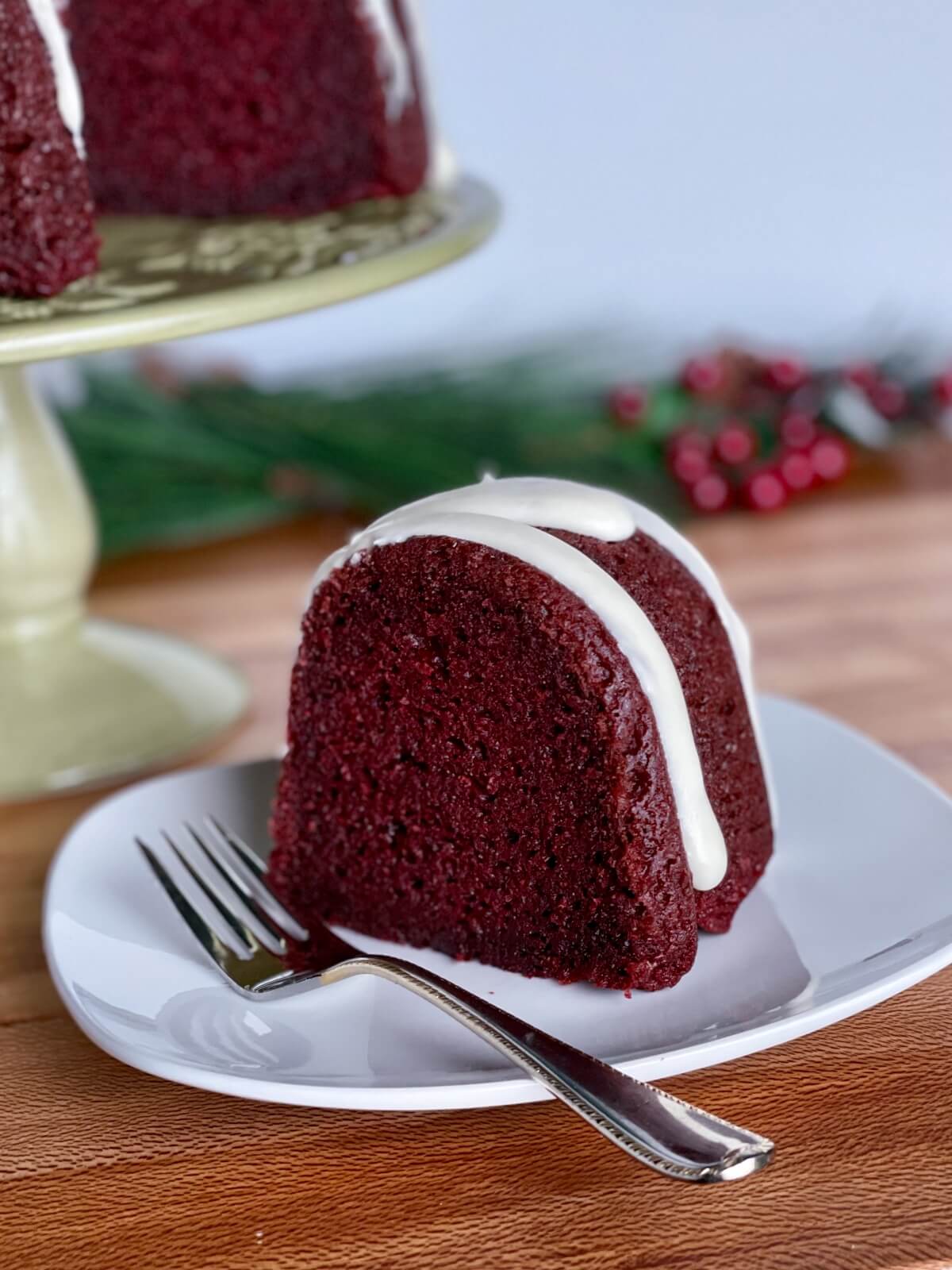 A closeup of a red velvet pound cake slice, on a plate with cream cheese icing and some holly berries and Christmas greenery in the background
