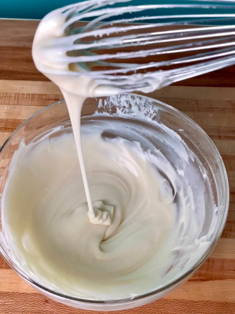 A bowl of cream cheese icing glaze with a whisk drizzling some of the icing into the bowl