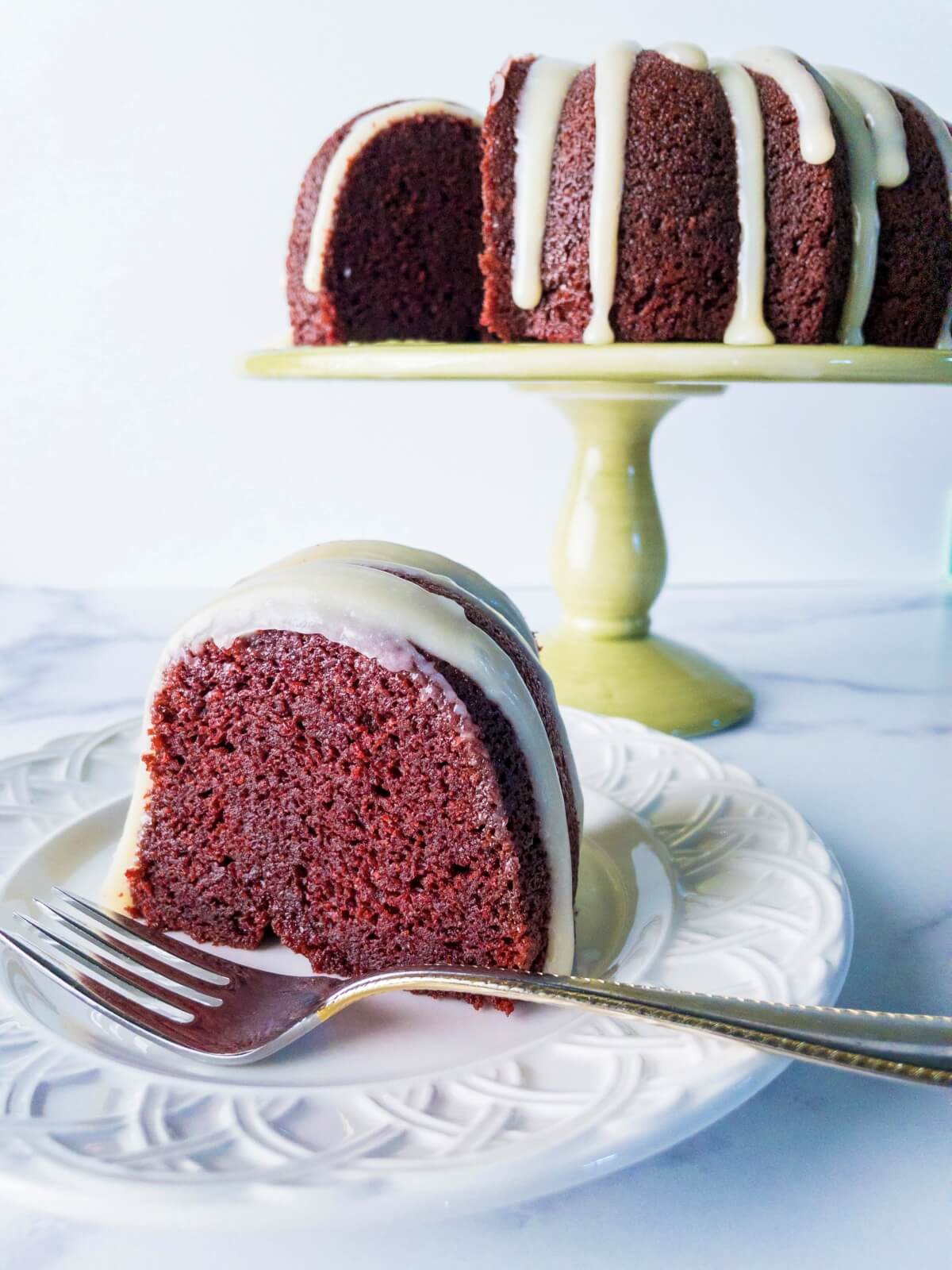 A slice of moist red velvet pound cake with a cake pedistal and the remaining cake in the background