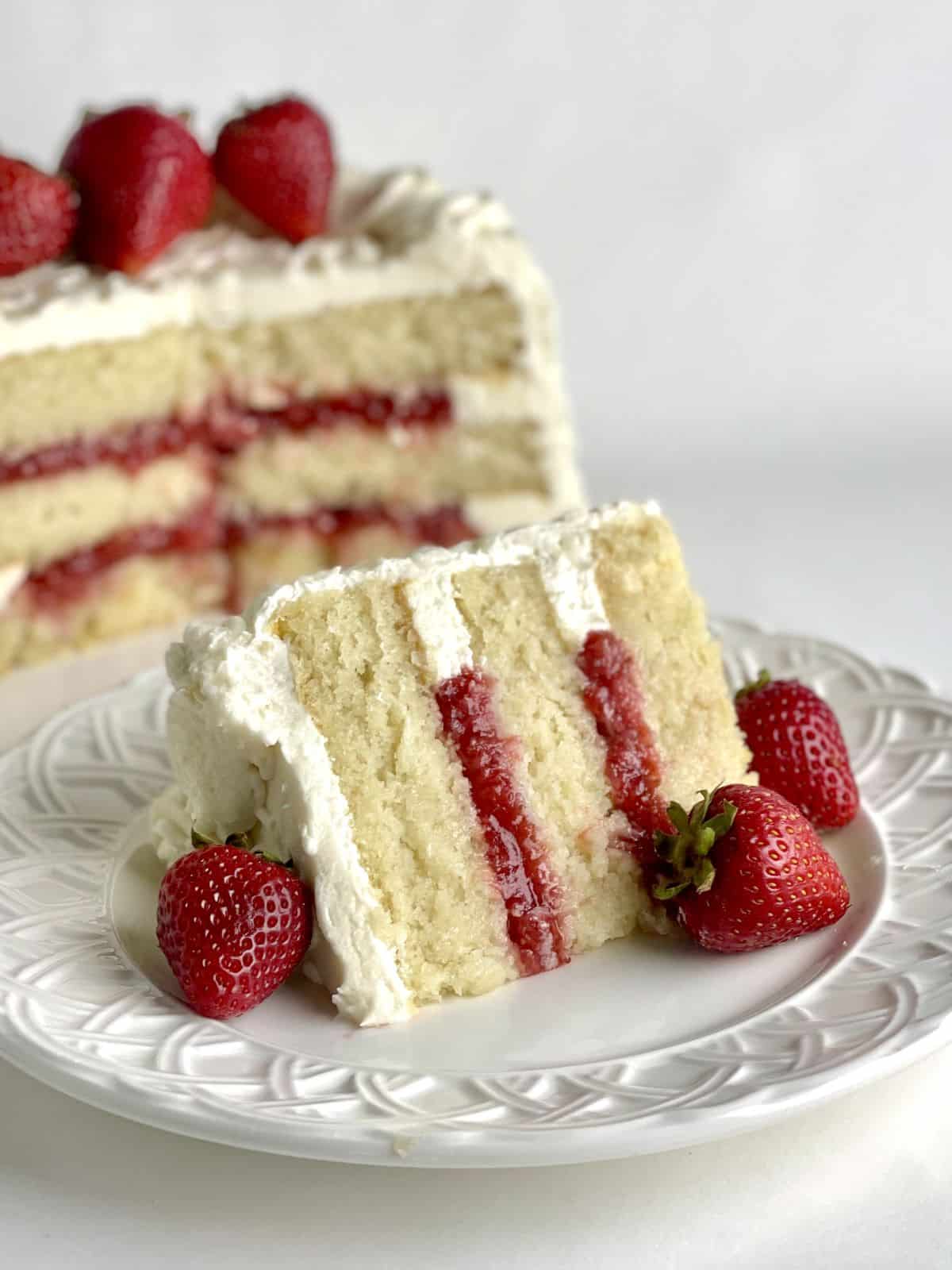 A slice of white cake with strawberry filling on a plate with the layer cake in the background.