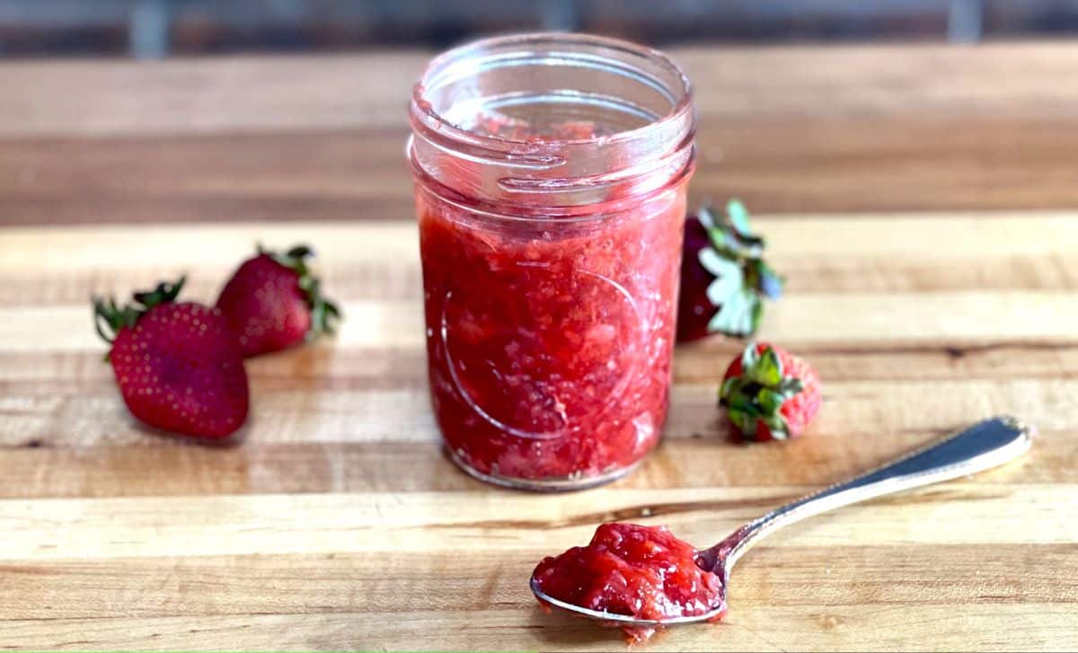 A jar of strawberry compote cake filling with a spoon and some fresh strawberries on a table.