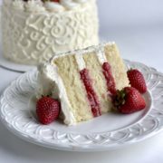A slice of vanilla strawberry cake with the decorated vanilla cake in the background.