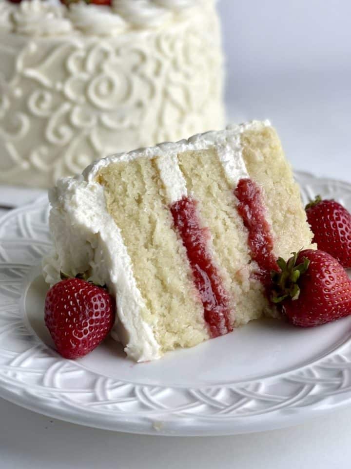 A slice of vanilla strawberry cake with the decorated vanilla cake in the background.