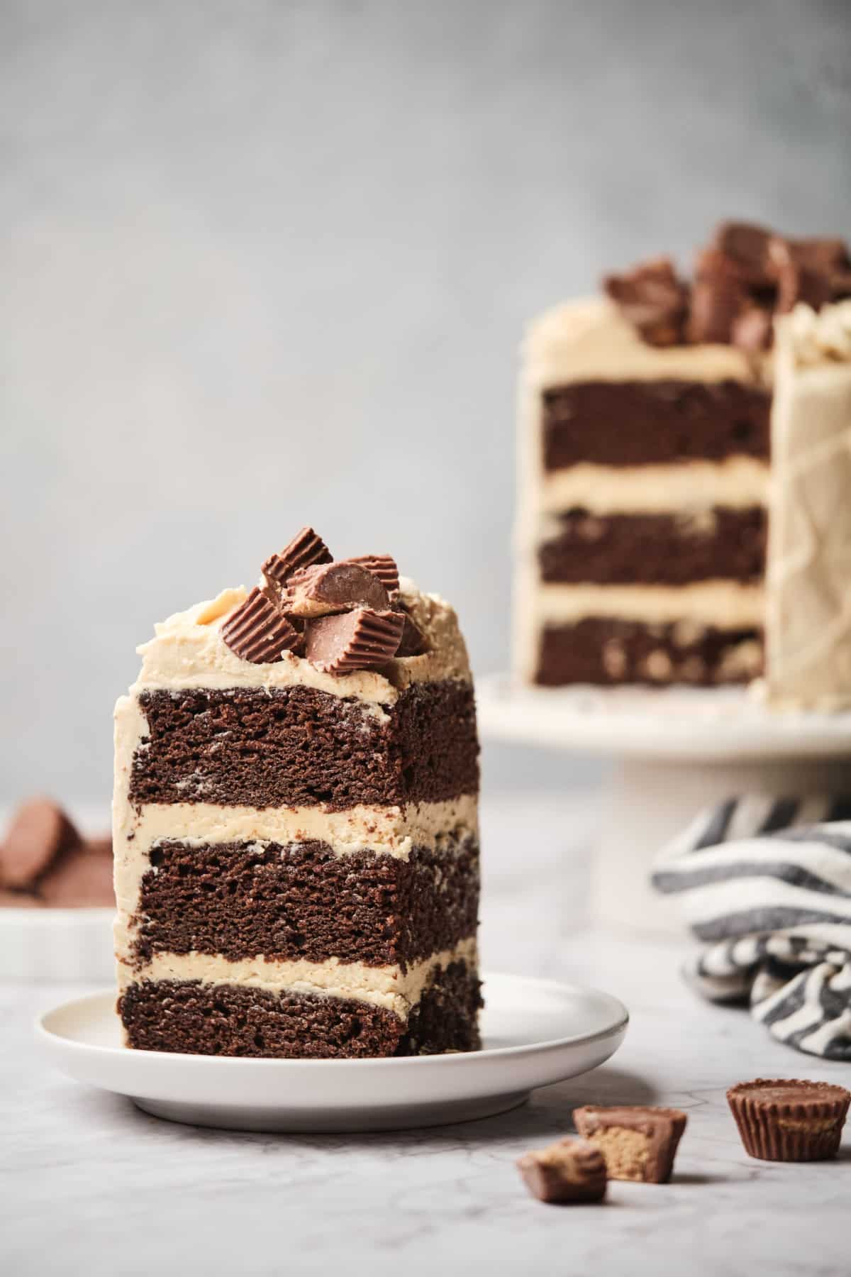 A slice of 3-layer chocolate cake with peanut butter frosting on a plate with the layer cake in the background.
