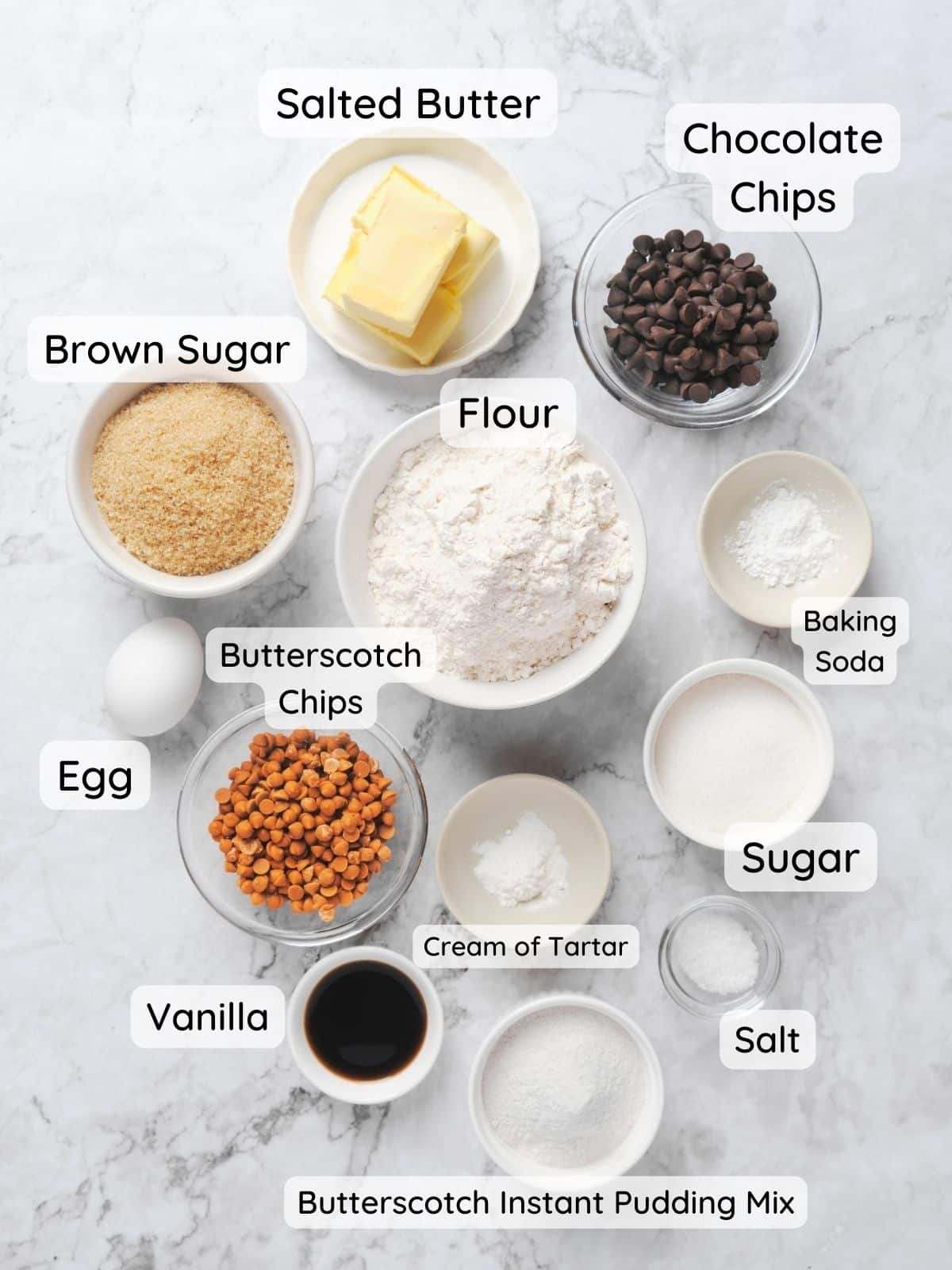 An overhead shot of chocolate chip butterscotch cookie ingredients with the text "flour, sugar, brown sugar, cream of tartar, baking soda, butterscotch chips, chocolate chips, vanilla, egg, butterscotch instant pudding mix, salted butter."