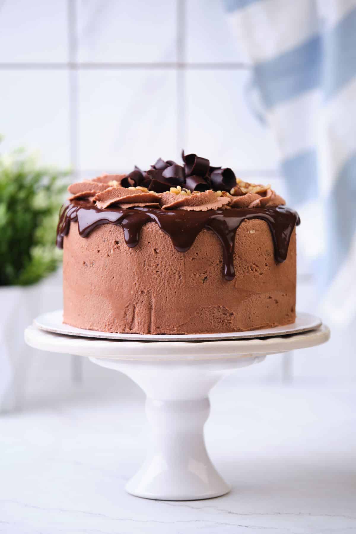 A chocolate cake on a cake stand decorated with chocolate drip and walnuts and chocolate curls.