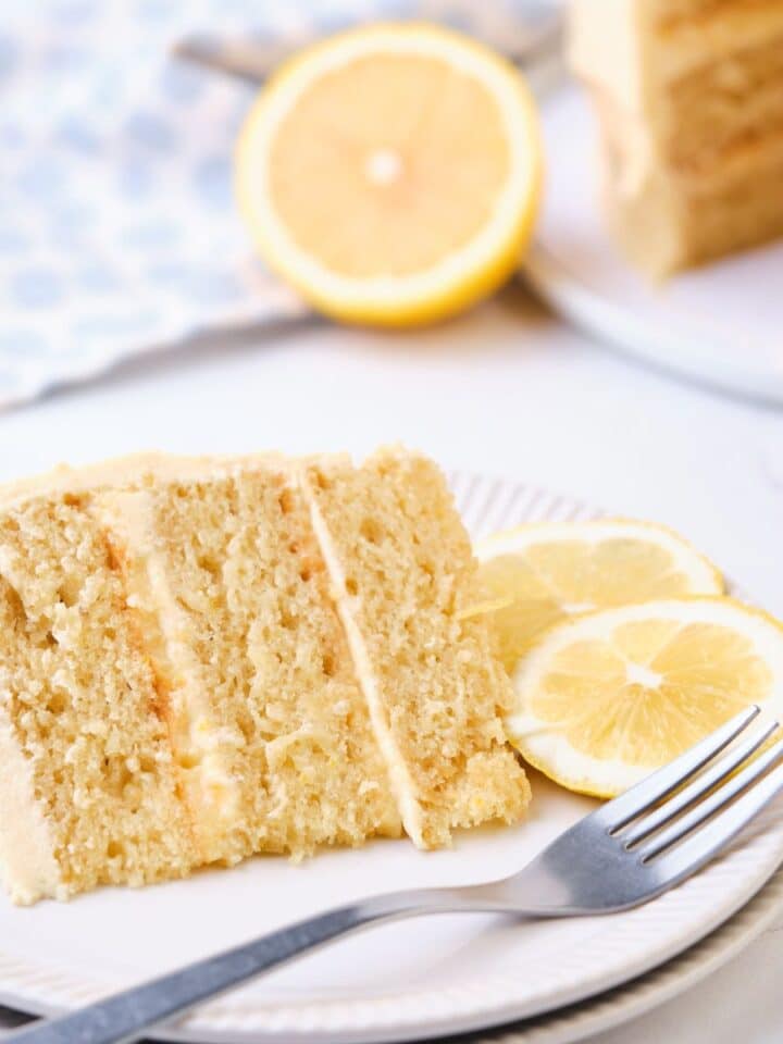 A slice of lemon curd cake on a plate with the lemon curd layer cake in the background.