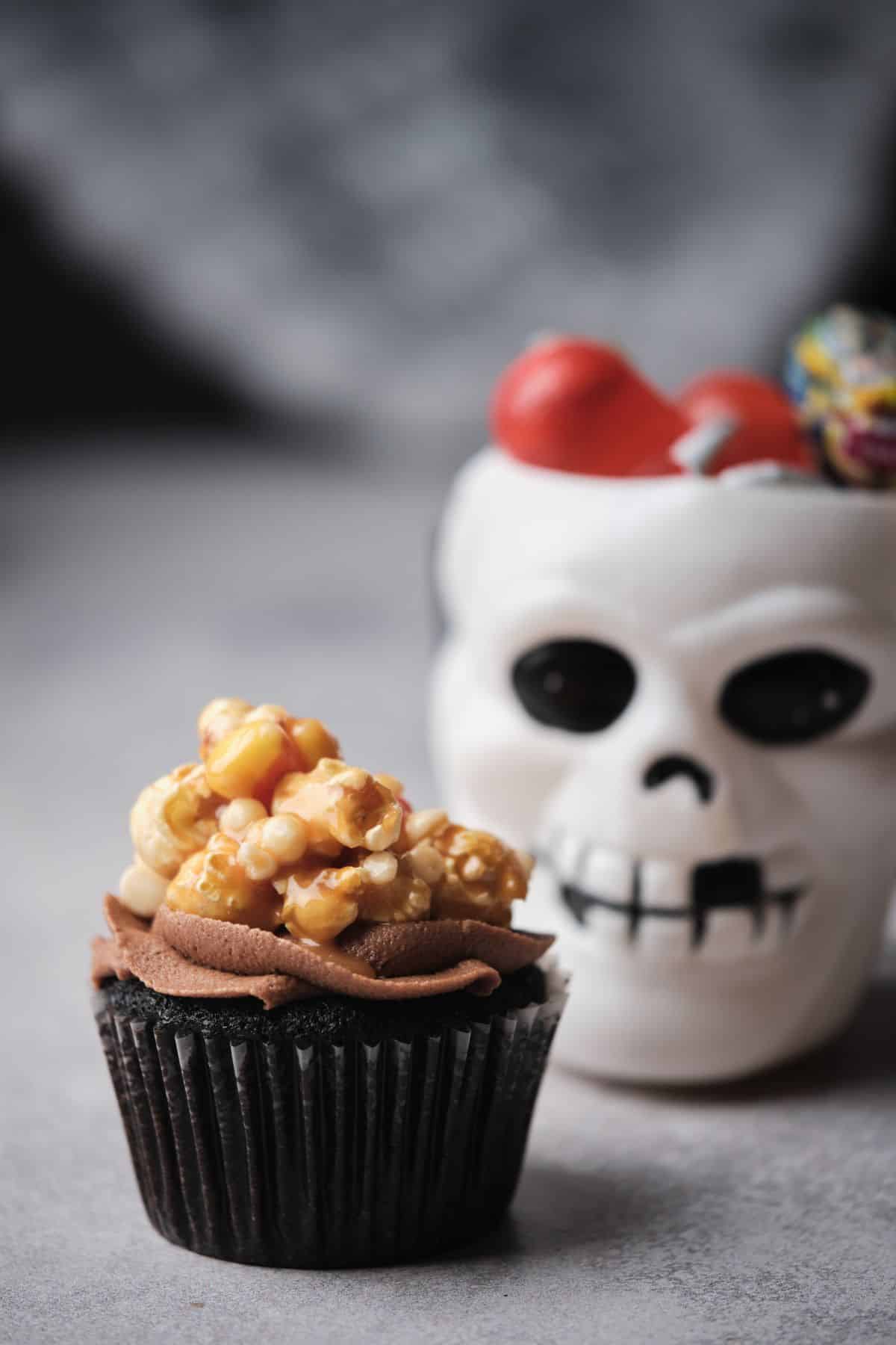 A Halloween Caramel filled chocolate cupcake topped with Caramel corn and Halloween decorations in the background.