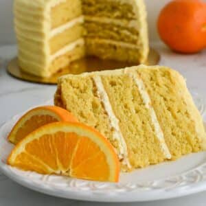 slice of moist orange almond cake on a white plate in front of a three layered cake.