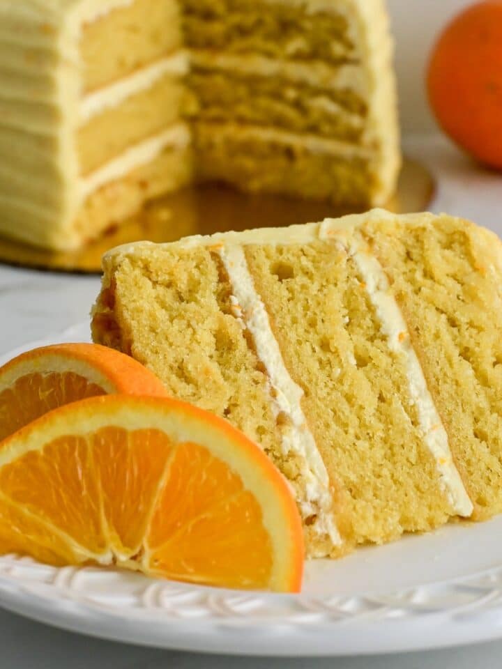 slice of moist orange almond cake on a white plate in front of a three layered cake.