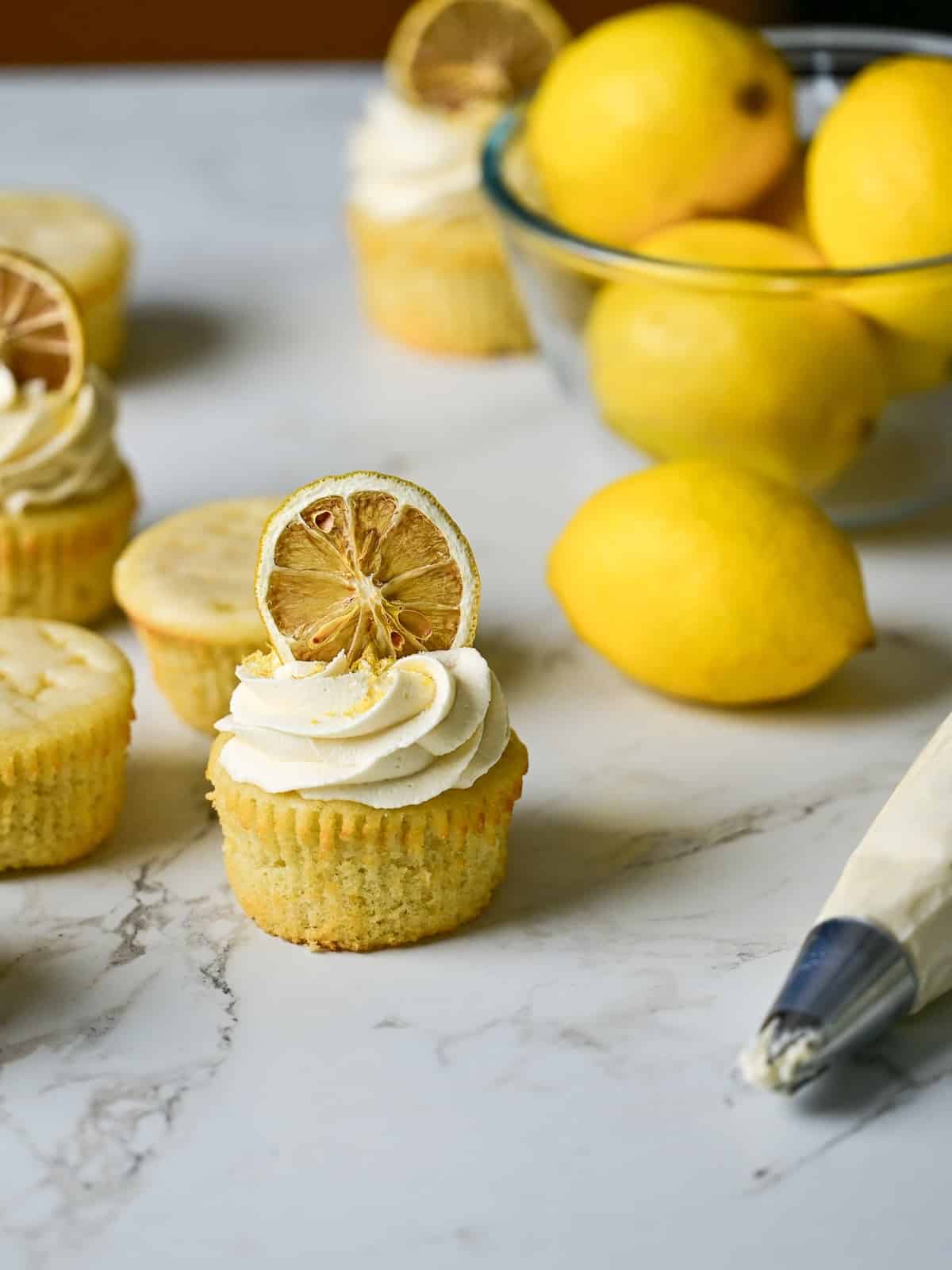 marble table with freshly made lemon drizzle cupcakes topped with frosting and dried lemon wheels.