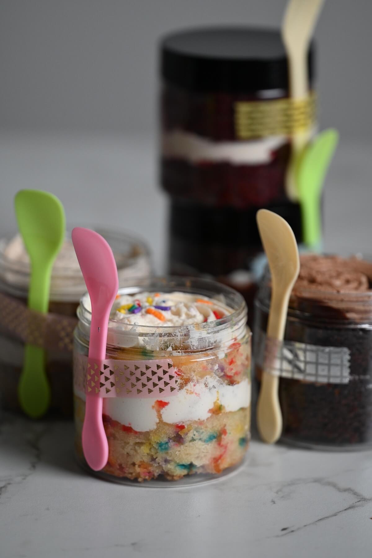 close up view of plastic jars with cake in the jar including funfetti and chocolate with attached sample spoons.