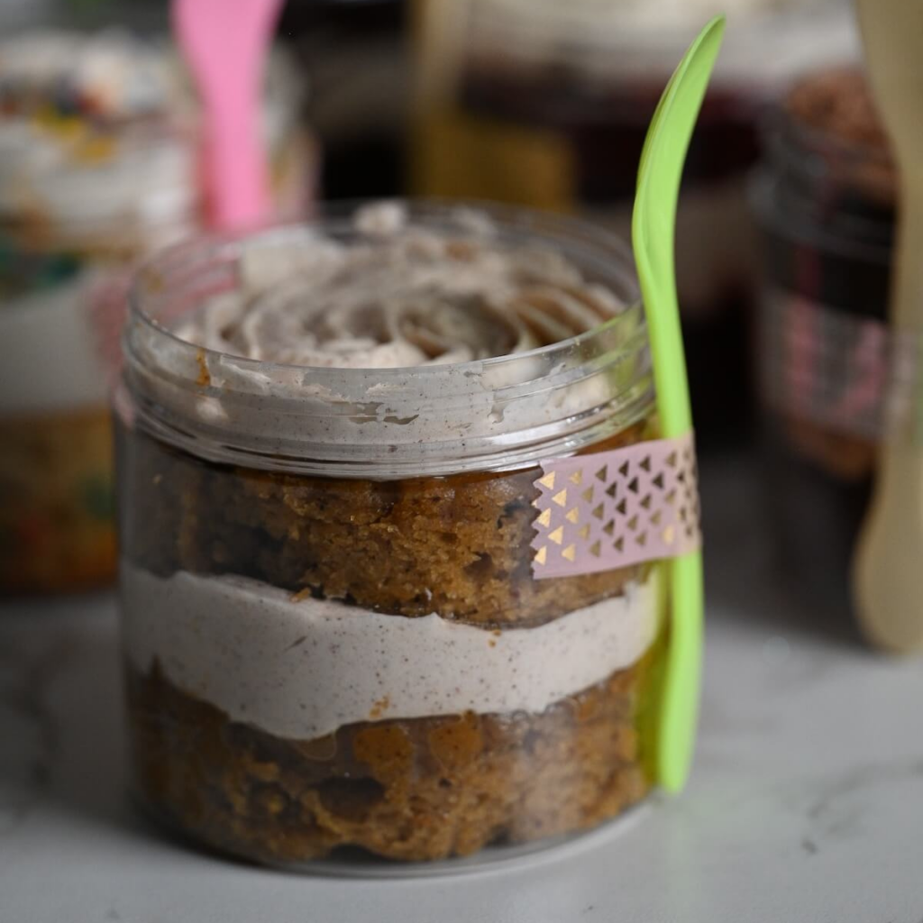 close up view of a plastic jar with layers of moist carrot cake and cinnamon buttercream with a spoon and washi tape attached to the jar.