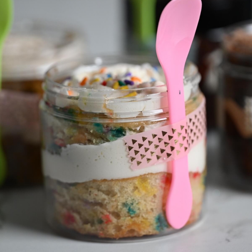 close up view of a plastic jar with funfetti cake, vanilla frosting, and rainbow sprinkles ready to serve.