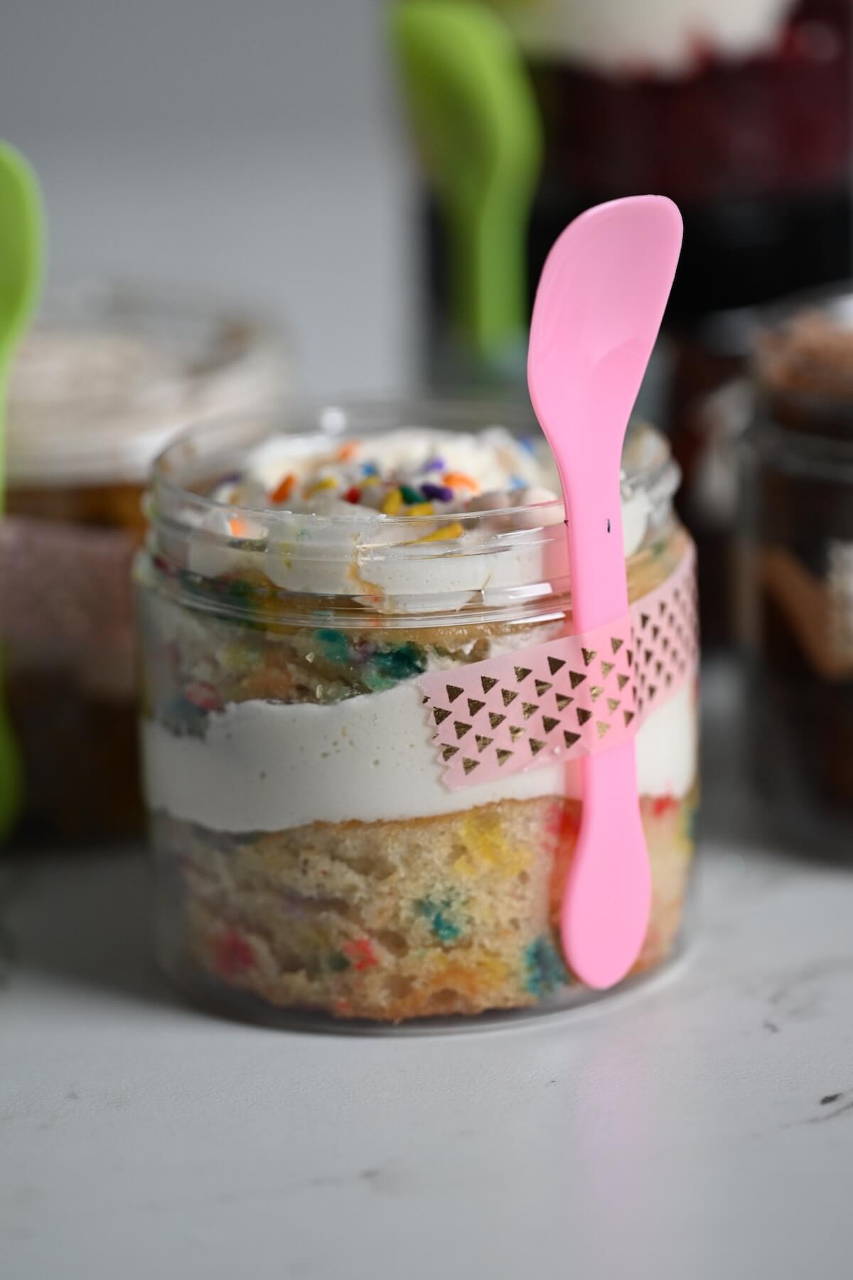 close up view of a cake in a jar with funfetti cake, vanilla buttercream, and a pink spoon taped to the jar with washi tape.