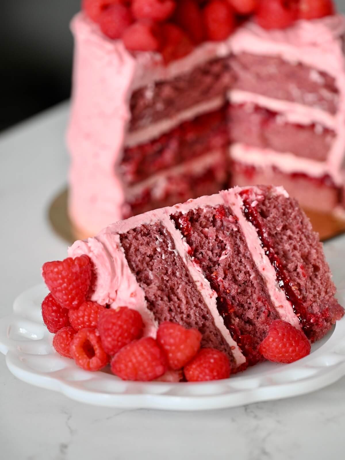 A raspberry layer cake with a slice on a plate and fresh raspberries.