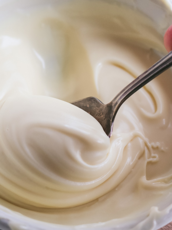 close up view of a spoon scooping up thick white chocolate ganache in a white bowl.