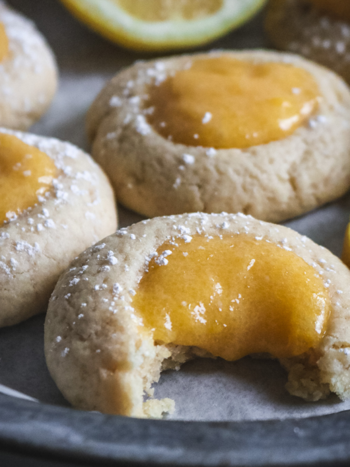 close up view of lemon curd thumbprint cookies on a metal tray with a bite taken out of one of them.