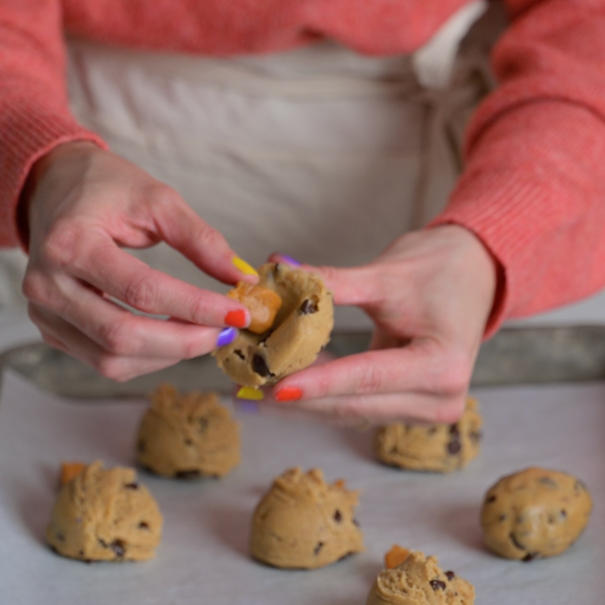 Hands stuffing a caramel candy into chocolate chip cookie dough balls on a baking sheet for gooey caramel-filled Halloween cookies.