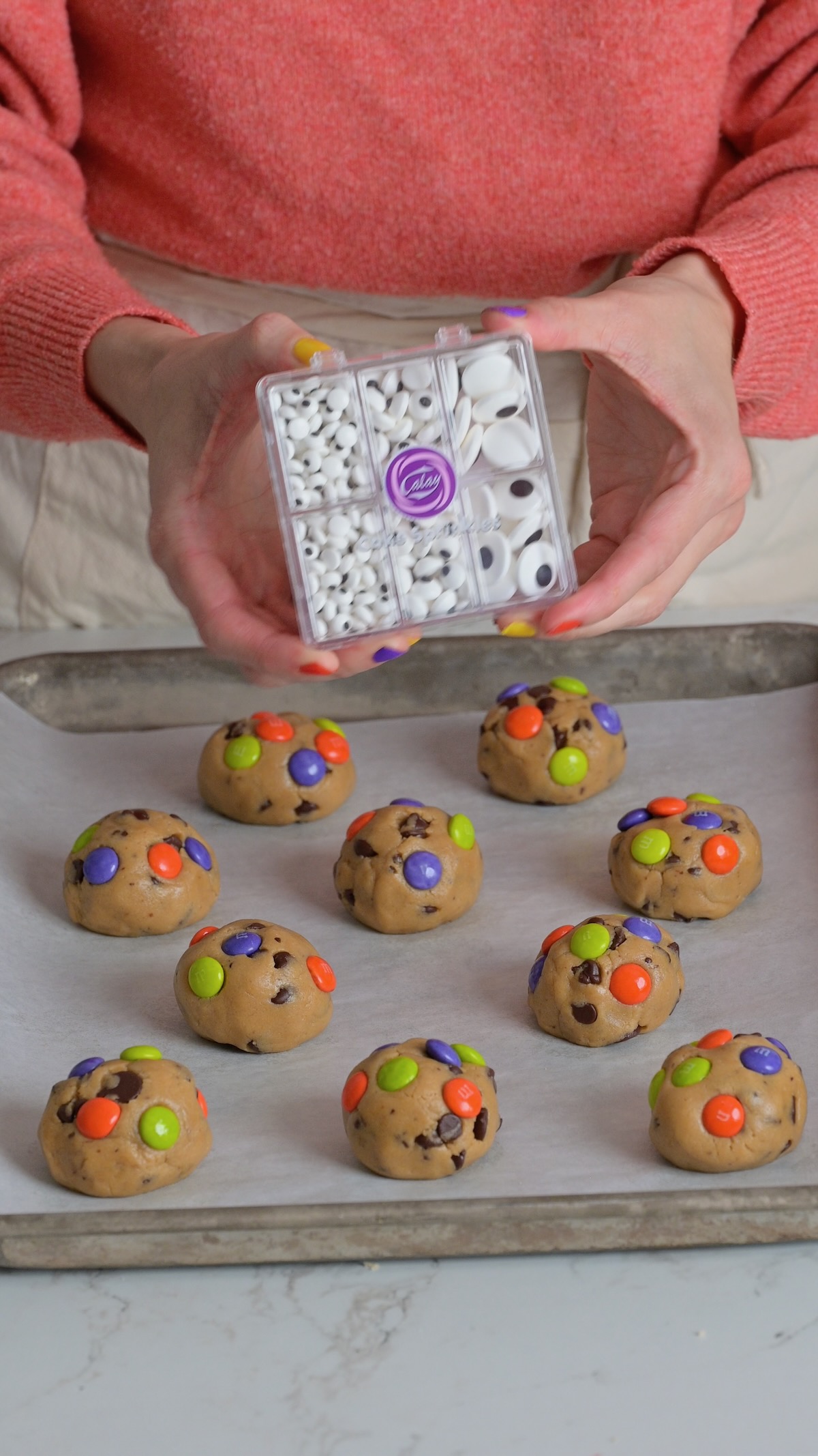 Hands holding a container of edible candy eyes above unbaked cookie dough balls decorated with Halloween M&M’s.