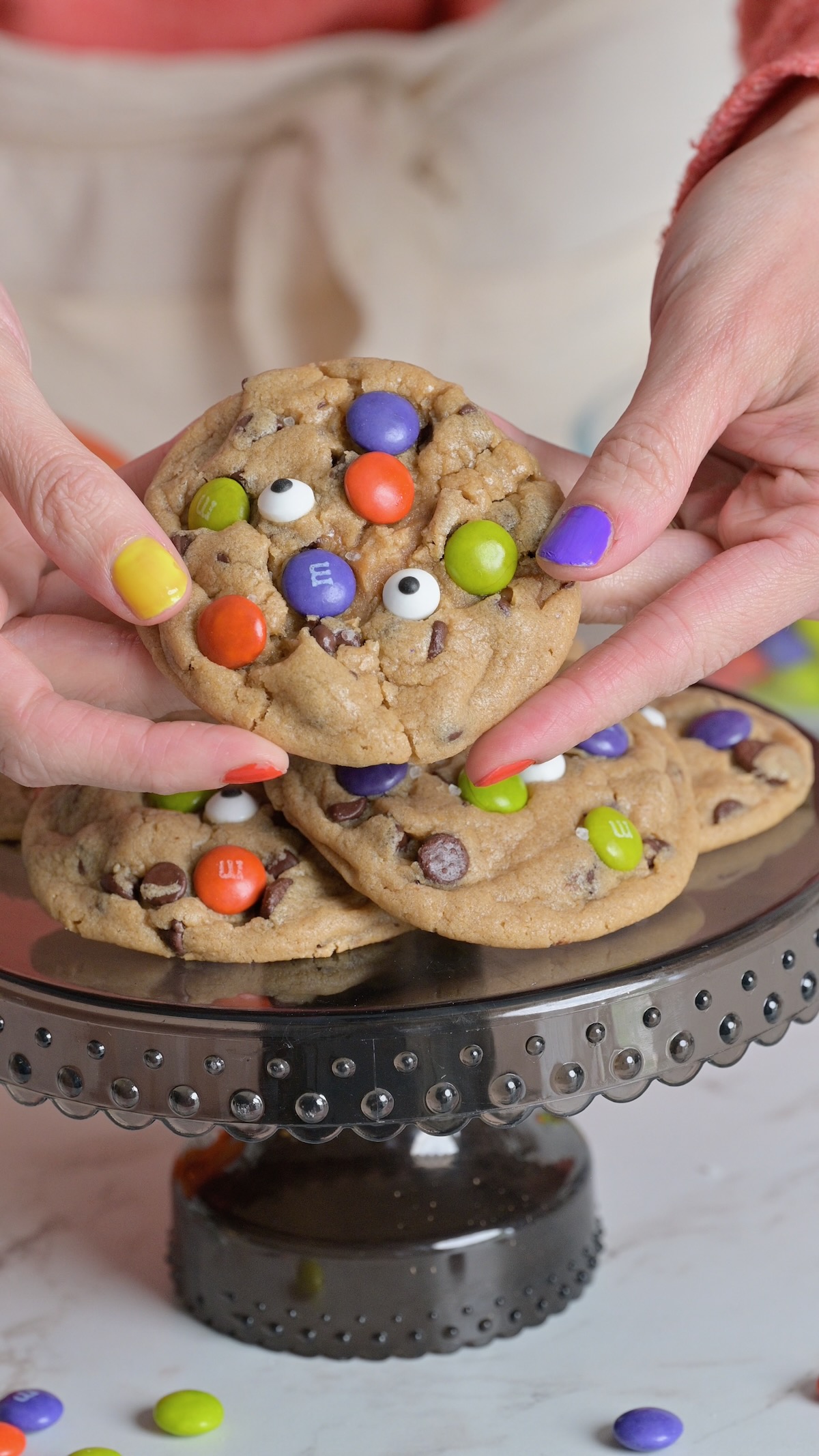 Close-up of hands holding a soft Halloween chocolate chip cookie filled with caramel and decorated with candy eyeballs and colorful M&Ms.