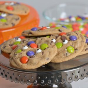 Baked Halloween chocolate chip cookies decorated with colorful M&M’s and candy eyeballs on a black cake stand.