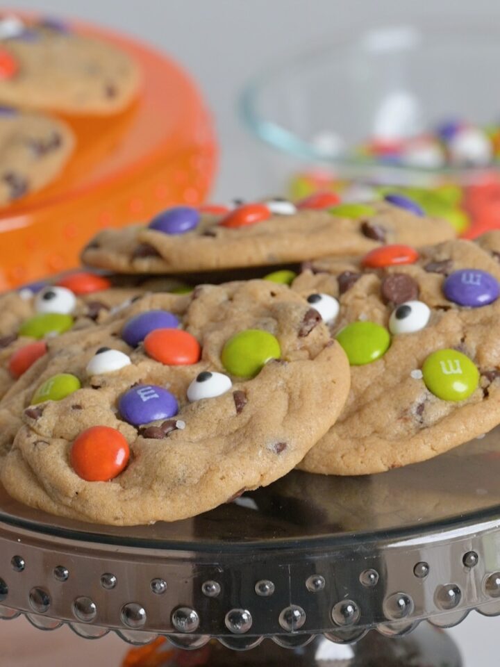 Baked Halloween chocolate chip cookies decorated with colorful M&M's and candy eyeballs on a black cake stand.