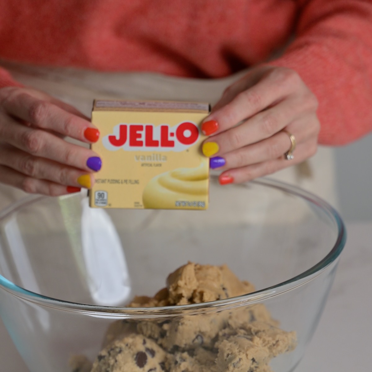 Hands holding a box of Jell-O vanilla pudding mix over a bowl of chocolate chip cookie dough.
